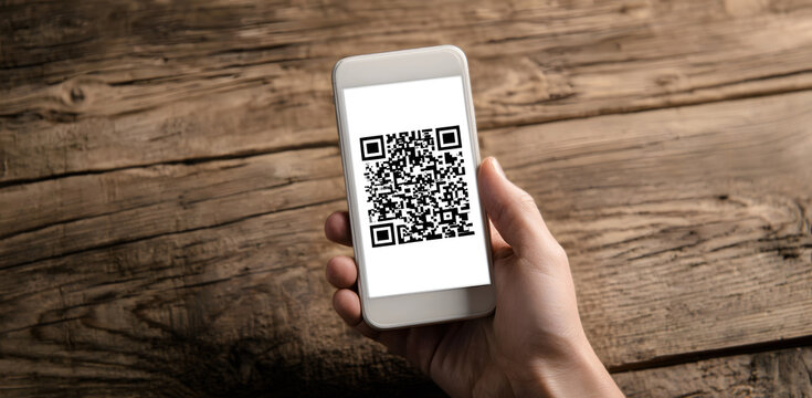 hand holding white smartphone with qr code visible on its screen placed against wooden background, symbolizing modern digital technology, contactless payment and secure mobile transactions