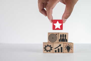 Hand placing a red star block on top of a pyramid of wooden blocks with business strategy icons (target, team, idea, growth, process), symbolizing the final step to success,quality,achieving goals.