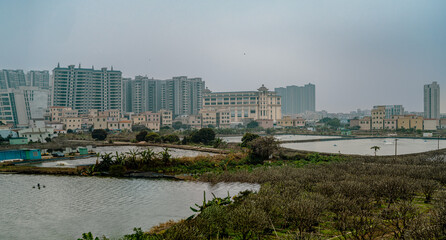 Fototapeta premium Between Guangzhou, Canton, and Macao, China, 3 March 2025. Panoramic view from a moving train across the Pearl River Delta showing rural ponds and new residential towers under cloudy sky.