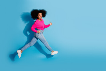 Obraz premium Cheerful young woman jumping energetically in pink sweater and blue jeans against a vibrant blue background showcasing joy