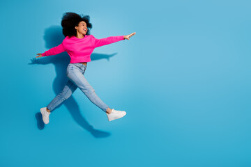 Fototapeta premium Cheerful young woman jumping against vibrant blue backdrop in stylish casual outfit showcasing lively and energetic pose