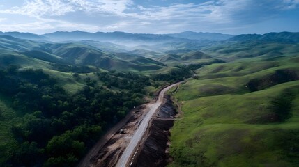 Fototapeta premium Aerial view of a new road under construction cutting through lush green rolling hills with distant misty mountains