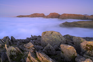 Bowfell summit rocks with incredible view looking over to Scafell Massif mountain range with a thick cloud inversion below. Lake District, UK.