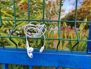Tangled White Wired Earbuds with Lightning Connector, Hanging on a Green Metal Mesh Fence Above a Blue Rail, Against a Backdrop of Green Foliage.