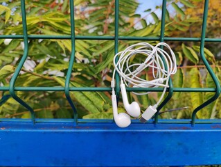 Tangled White Wired Earbuds with Lightning Connector, Hanging on a Green Metal Mesh Fence Above a Blue Rail, Against a Backdrop of Green Foliage.
