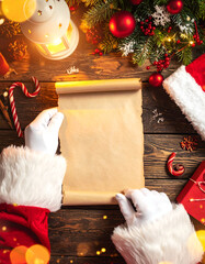 Top view of Santa&rsquo;s hands with empty wish list paper among festive holiday ornaments and presents.