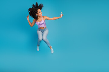 Obraz premium Young stylish woman jumps in mid air against blue background wearing purple top jeans and sneakers conveying joy energy and fashion lifestyle