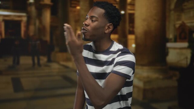 Man wearing striped shirt blows kiss with open palm gesture in church hall amid stone columns; devotion.