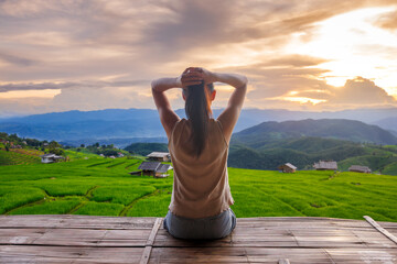 a young woman sitting on a bamboo terrace overlooking lush green rice fields at Ban Pa Bong Piang in Chiang Mai, relaxing with hands behind her head while watching a peaceful sunset over the mountain 