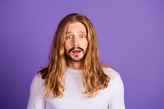 Young handsome man with long blond hair in a white shirt poses against a purple background with a surprised expression