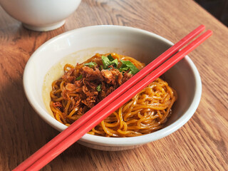A close-up of a bowl of Indonesian-style fried noodles topped with crispy shallots and fresh herbs, served on a wooden table with red chopstick in warm natural light.