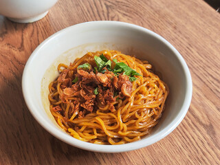 A close-up of a bowl of Indonesian-style fried noodles topped with crispy shallots and fresh herbs, served on a wooden table in warm natural light.