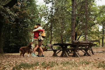 Young couple hugging in a forest clearing with two dogs nearby, surrounded by wooden benches. Concept of love, friendship, and hiking with pets