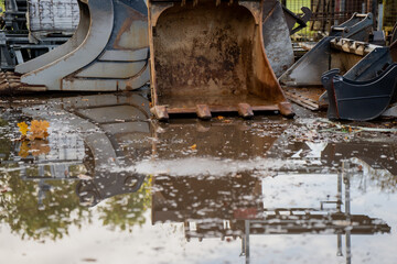 Fototapeta premium Rusty excavator bucket standing beside other digging attachments in wet construction yard with puddle reflection, showing durability, engineering precision, and equipment readiness for excavation work