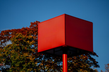 Red cube-shaped billboard for advertising pole against blue sky and autumn trees, showing geometric...