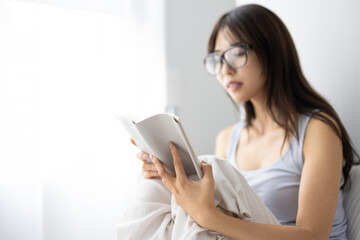 Fototapeta premium Young Woman Reading a Book on Bed with Glasses in Natural Light