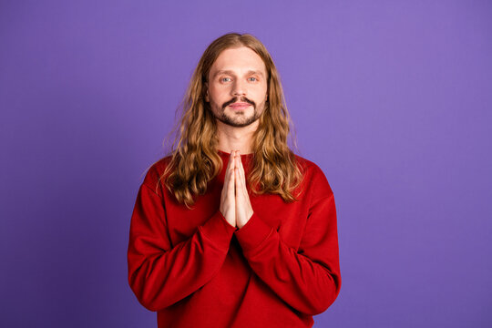 Young man with long hair in a red sweater stands with hands pressed together against purple background - Powered by Adobe