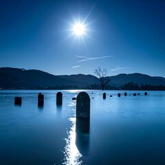 Serene nightscape features a bright celestial body illuminating a lake with submerged posts, mountains, and lone tree