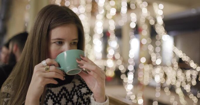Young woman enjoying a warm cup of coffee in a festive setting with sparkling lights and a relaxed atmosphere