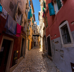 Obraz premium Rovinj, Croatia - August 15, 2025: Narrow Alley in Rovinj – colorful old buildings with open shutters and laundry hanging across a sunlit cobblestone street in the charming old town of Rovinj, Croatia