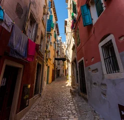Fotobehang Smal steegje Rovinj, Croatia - August 15, 2025: Narrow Alley in Rovinj – colorful old buildings with open shutters and laundry hanging across a sunlit cobblestone street in the charming old town of Rovinj, Croatia  © Алексей Горовой