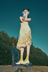 Young woman in yellow dress standing playfully on transparent chair outdoors.