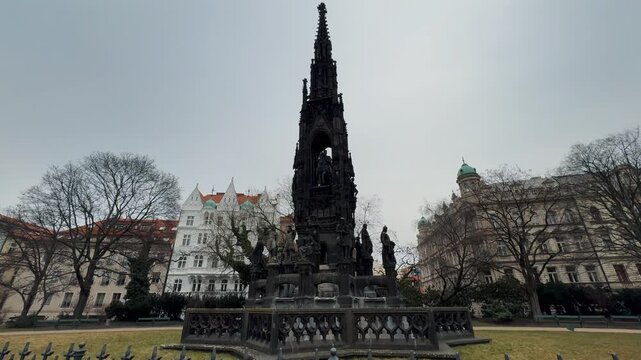 The dramatic Gothic Revival Kranner Fountain  stands tall in a central Prague park, featuring intricate ironwork and heroic statues under a cloudy sky.

