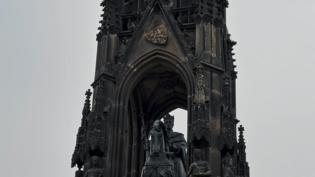 The dramatic Gothic Revival Kranner Fountain  stands tall in a central Prague park, featuring intricate ironwork and heroic statues under a cloudy sky.

