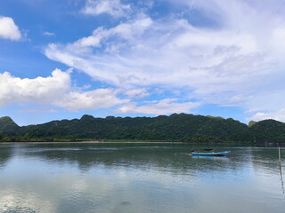 A view of a wide river and mountains covered with large greentrees. Sky, river, clouds, and a long-tailed boat moored in the middle of the river for fishing. Tranquility and natural beauty. 
