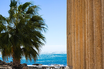 A Scenic View of Ancient Columns and Lush Palm Trees Overlooking a Tranquil Ocean Under a Bright Blue Sky