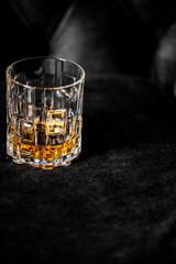 A luxurious crystal whiskey tumbler filled with golden brown liquor, set against a dark background. The glass features a geometric, textured design.