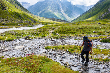 Naklejka premium Hiker crossing a stream surrounded by lush green valleys and towering mountains during a scenic trekking adventure.