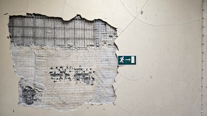 Cracked wall with exposed reinforcement mesh and a green emergency exit sign indicating direction © Egor Lint