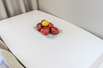 still life featuring a plate of red apples and a lemon on a white table. The background is a light, neutral color
