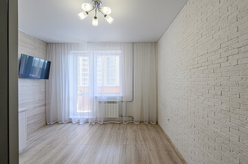 A bright, modern room featuring a TV, a window with sheer curtains, a light fixture, light wood flooring, and a white brick wall