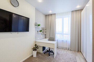 A modern room features a white desk with a gray chair near a window with curtains. A TV hangs on the wall, and there are shelves with decorative items