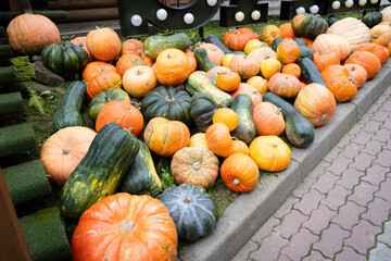A large, colorful pile of various pumpkins and squashes, including orange and dark green varieties, displayed outdoors next to a curb and a decorative sign with lights.