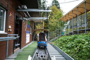 An elevated metal track structure of an alpine coaster (rodelbahn) featuring a looping turn, set on a grassy hill surrounded by a forest with autumn foliage under a cloudy sky.