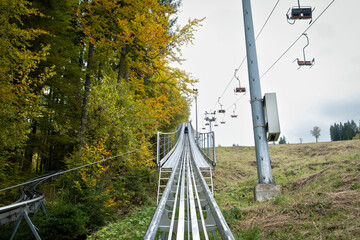 An elevated metal track structure of an alpine coaster (rodelbahn) featuring a looping turn, set on a grassy hill surrounded by a forest with autumn foliage under a cloudy sky.