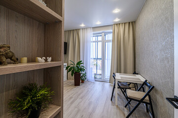 A cozy living space with light wood floors, a shelf unit, a dining table, and plants. Natural light floods the room through a large window