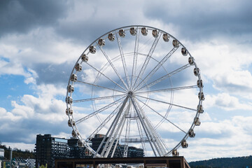 A large white Ferris wheel with enclosed gondolas stands tall against a dramatic sky with blue...