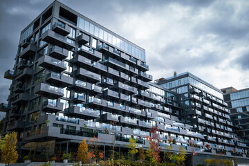 Modern, dark gray facade of a high-rise residential building with projecting balconies and large glass windows, set against a dramatic, cloudy sky, with a touch of autumn foliage. © Alisa