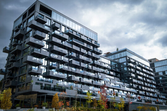 Modern, dark gray facade of a high-rise residential building with projecting balconies and large glass windows, set against a dramatic, cloudy sky, with a touch of autumn foliage.