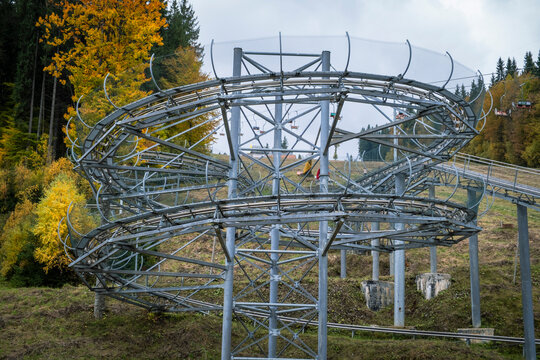 An elevated metal track structure of an alpine coaster (rodelbahn) featuring a looping turn, set on a grassy hill surrounded by a forest with autumn foliage under a cloudy sky.