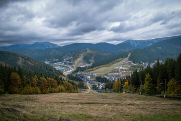 Wide panoramic view of a large mountain resort complex nestled in a valley with ski slopes and modern buildings, surrounded by dense forests and layered mountains under a dramatic cloudy sky in autumn