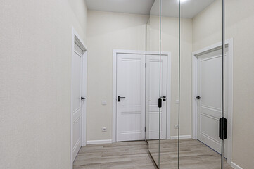 A bright hallway features white doors with black handles and a large mirror, reflecting the doors and light-toned walls