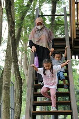 Obraz premium A caring mother wearing a hijab and mask helps two young children descend a wooden staircase in a lush park, conveying family safety, supervision, and outdoor time.