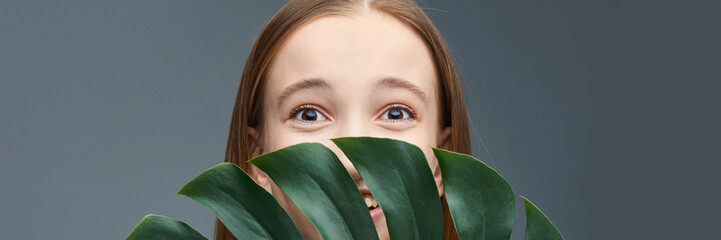 Young caucasian female peeking behind green leaves against gray background.
