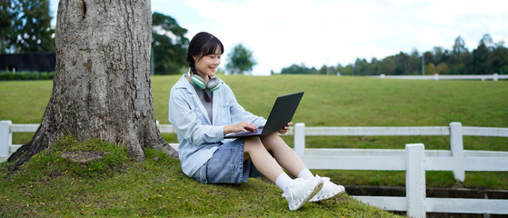 Fototapeta premium Concentrating young Asian freelancer working on a laptop while using a smartphone in a park, enjoying the fresh air and the beauty of nature in a serene outdoor setting