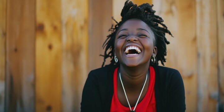 A woman with dreadlocks is smiling and laughing. She is wearing a red shirt and a necklace
